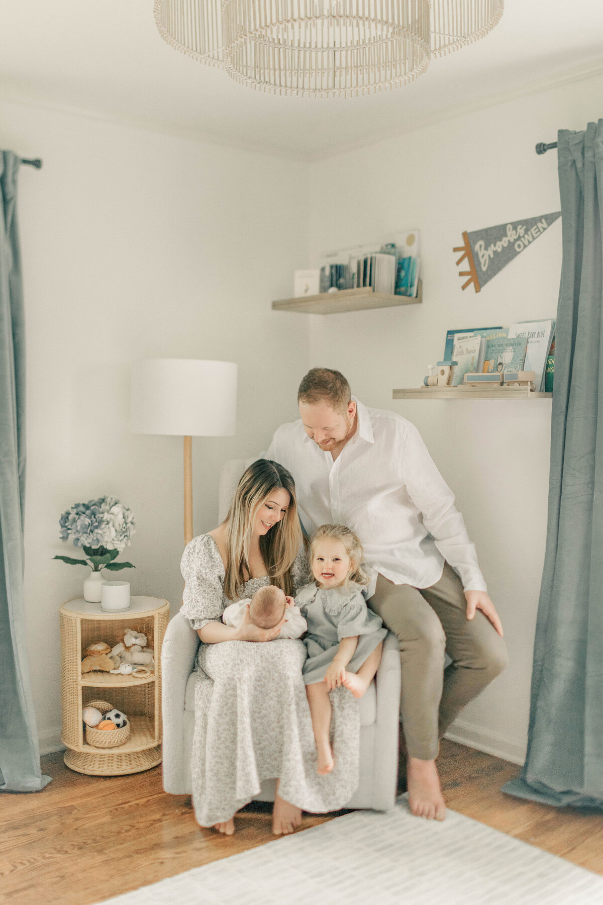 Family of four sitting closely in a nursery chair, with mom holding newborn and toddler on dad’s lap during an in-home newborn session in Kansas City.