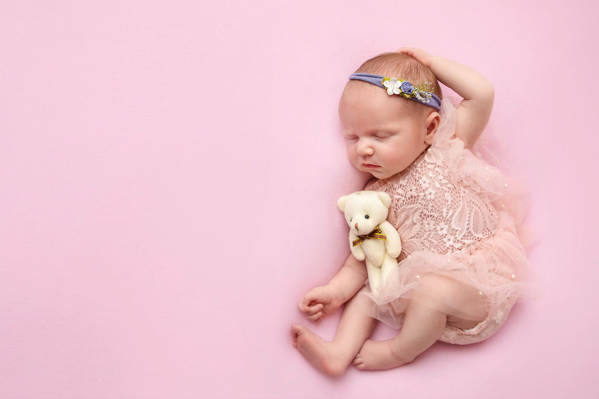Baby girl laying on a pink blanket with her hand on her head and holding a tiny teddy taken by Los Angeles newborn photographer Elsie Rose