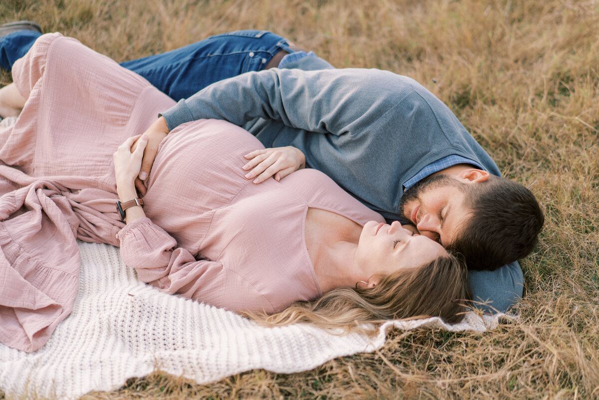 Man in a blue shirt and blue jeans lying next to a woman in a blush dress with their hands around her belly in a field.