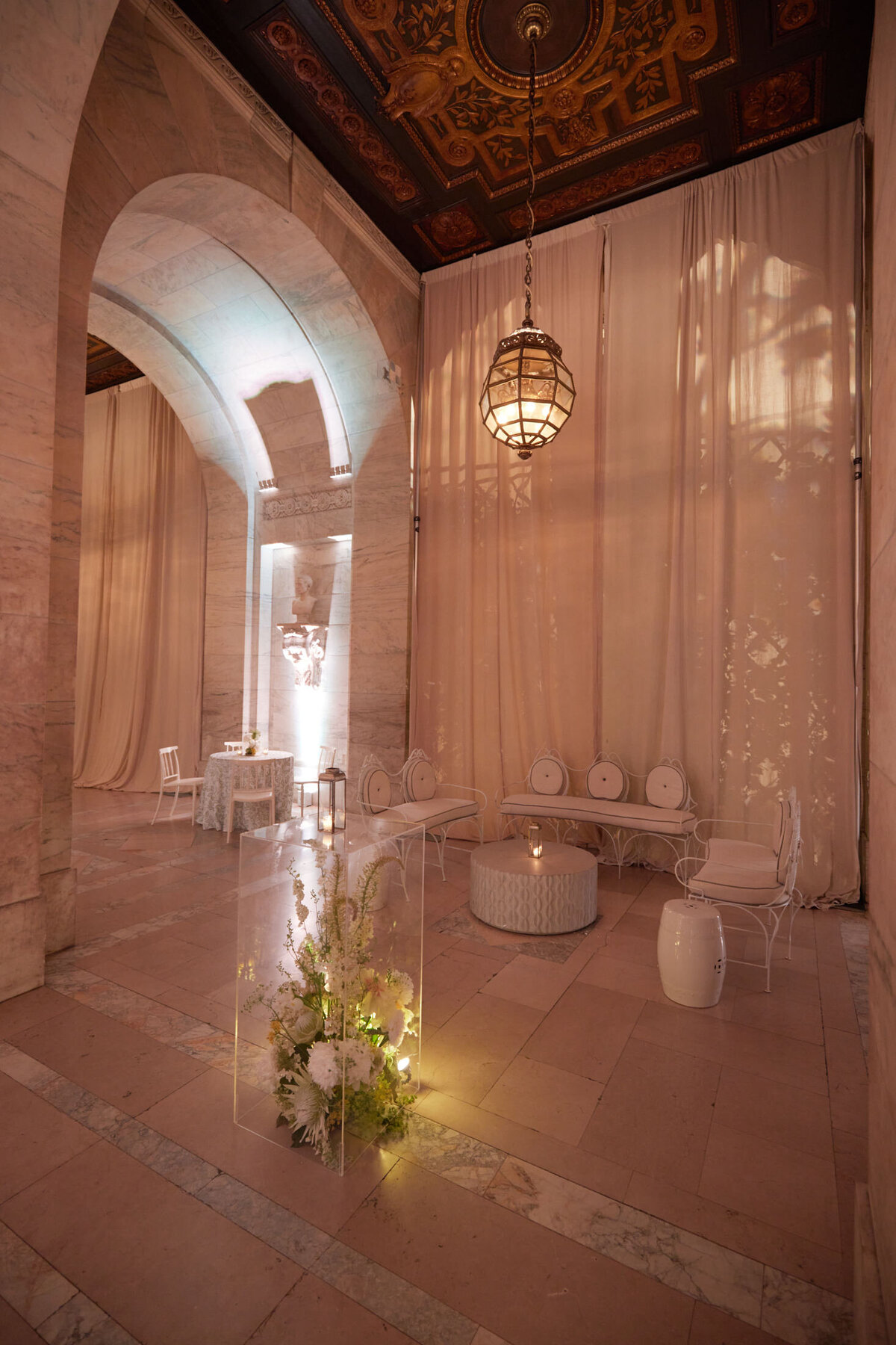 The photo was captured in New York City at The New York Public Library by photographer Shawn Connell. The luxurious, softly lit interior features tall, arched ceilings and marble walls, creating an elegant atmosphere. The space is tastefully decorated with draped curtains, a large hanging lantern, white furniture, and beautiful floral arrangements, adding to its charm. Additionally, a small round table and chairs are set in the background, offering a cozy nook within this beautiful setting. Videographers: Aaron Navak Films.