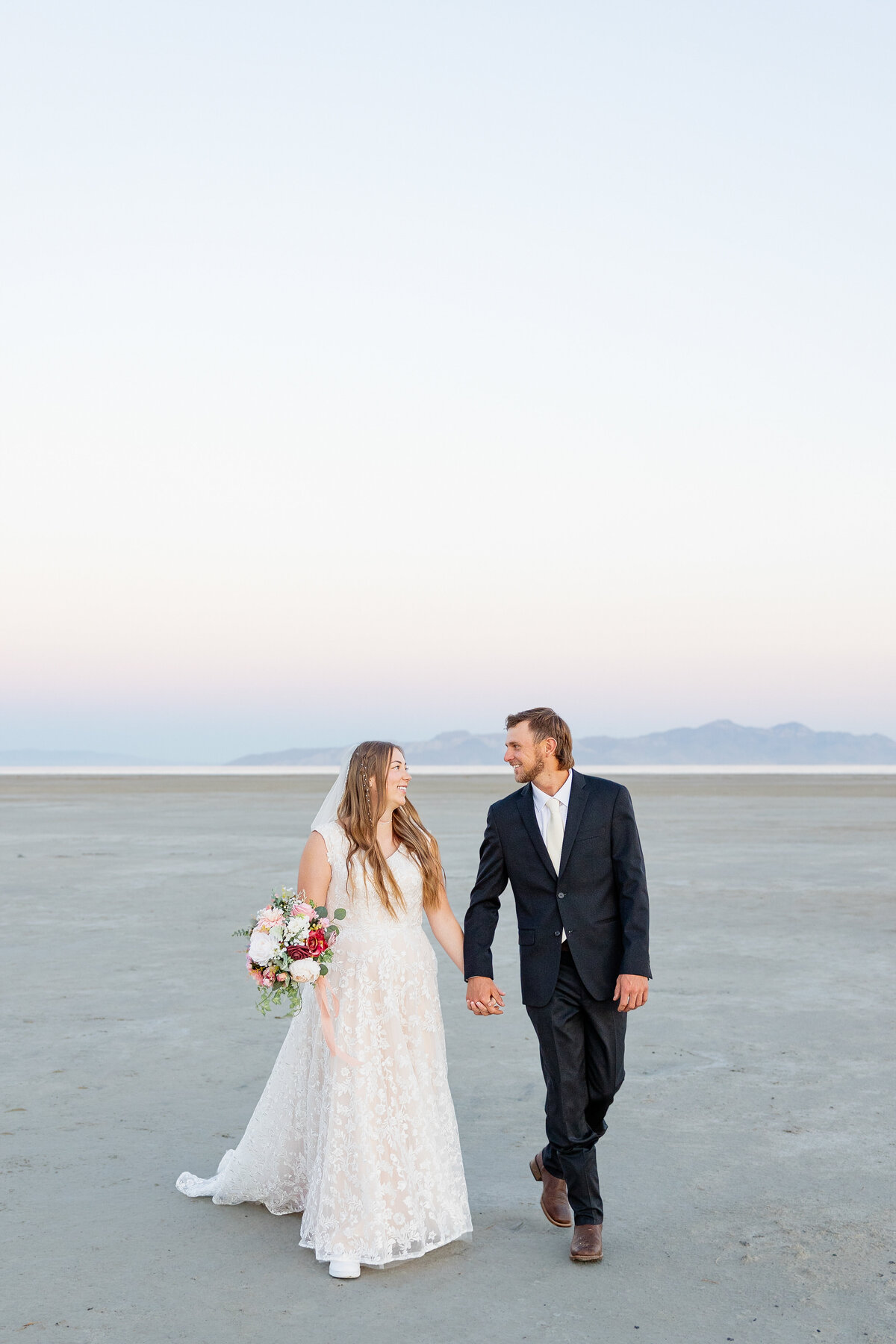 Bride in a white lace gown and groom in a black suit walk side by side, smiling at each other.
