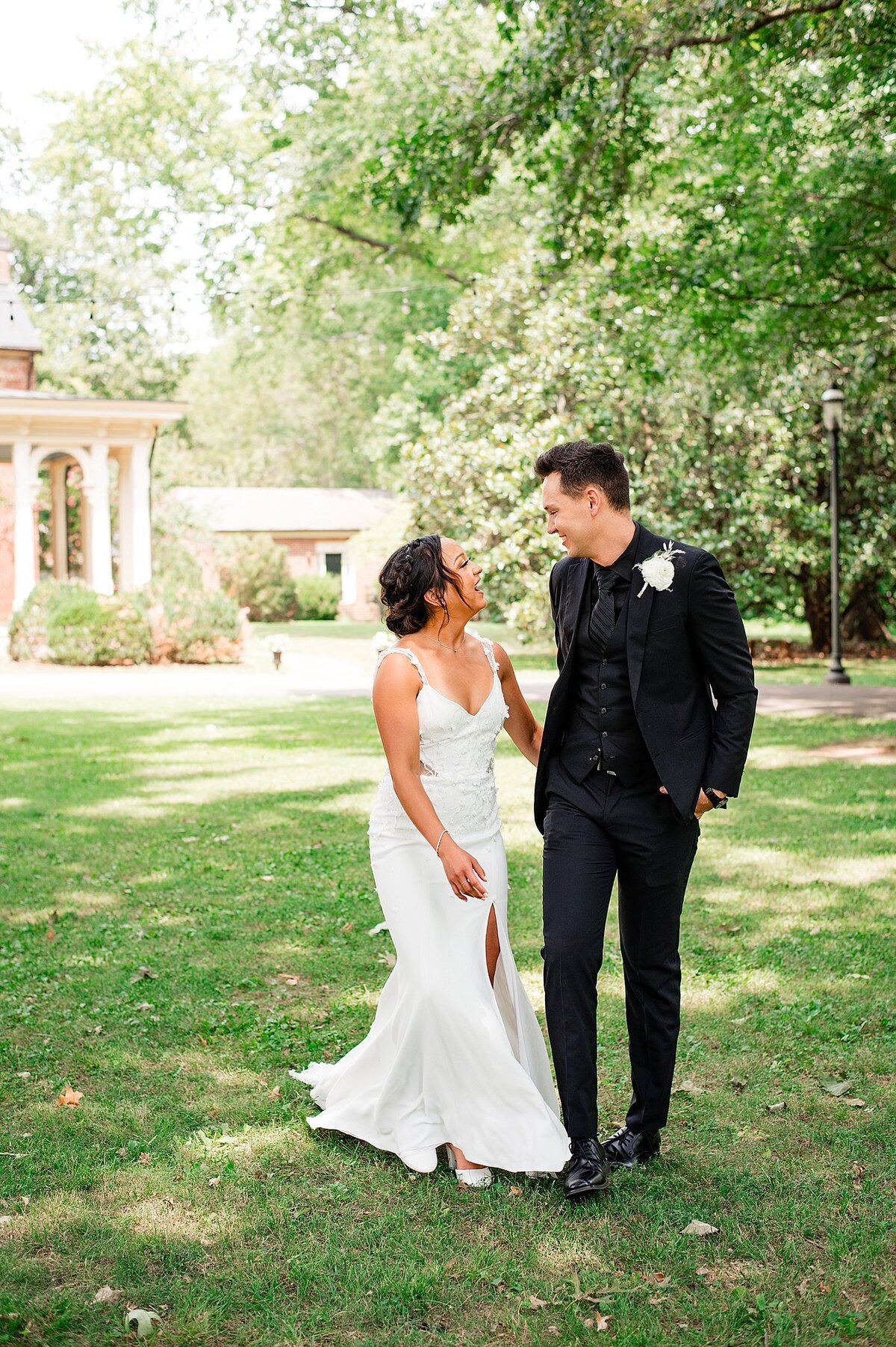 Bride wearing a fitted wedding gown with a flaired skirt and thigh high slit laughs up at the groom who is holding her hand. The groom is wearing a black suit with a black shirt and a white rose boutonniere. They are walking the grounds at Oaklands Mansion.