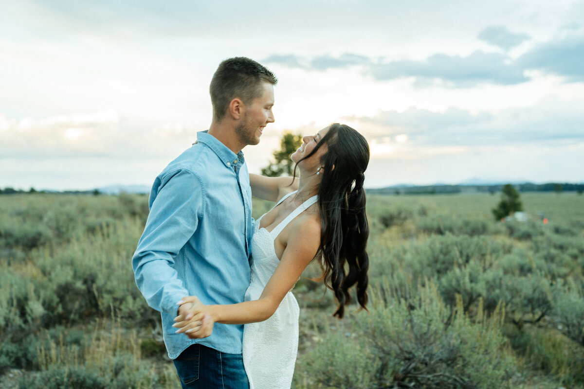 A bride and groom dance together outside in Grand Teton National Park. She has her hand on his shoulder and he has his hand on her waist. They're looking at each other, smiling big. Sagebrush is behind them.