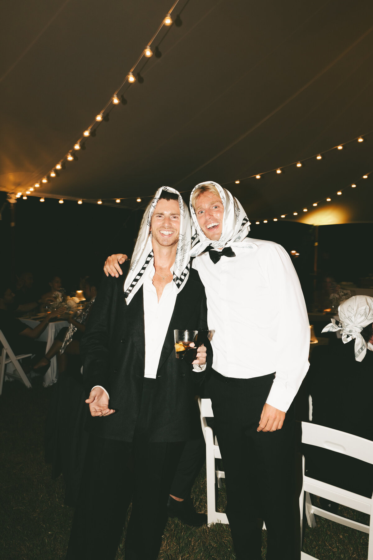 Two wedding guests wearing scarves on their heads laugh and pose under string lights at a tented wedding reception.