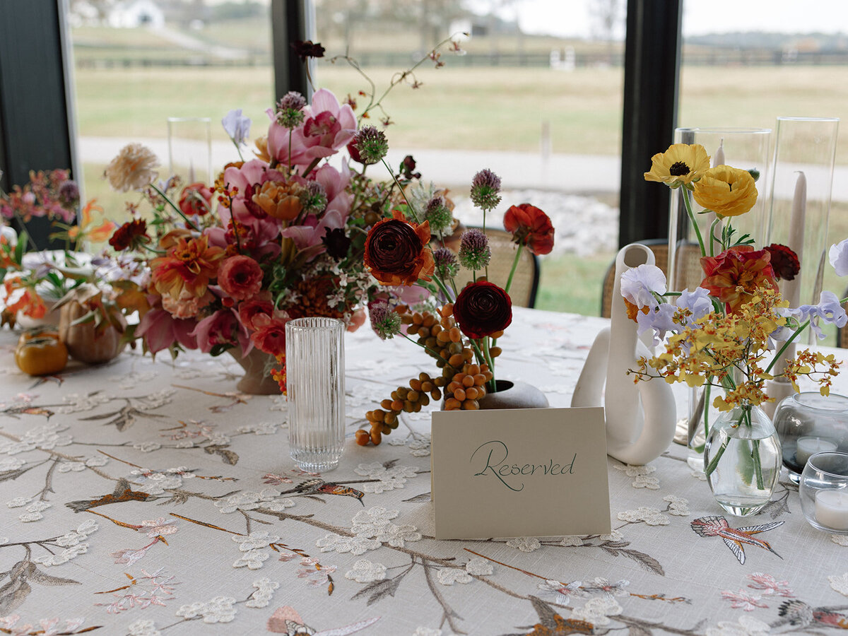 Clustered and styled floral arrangements, fruits, and art pieces for fall wedding reception. Low centerpieces mixed with ikebana arrangements and bud vases decorate the reception tables at this fall wedding. Floral centerpieces featuring orchids, ranunculus, sweet peas, and bittersweet. Styled wedding tables with fruits, curated candles, and floral arrangements. Tasteful fall color palette of copper, ruby, gold, burgundy, rust, and lavender. Design by Rosemary & Finch Floral Design in Nashville, TN.