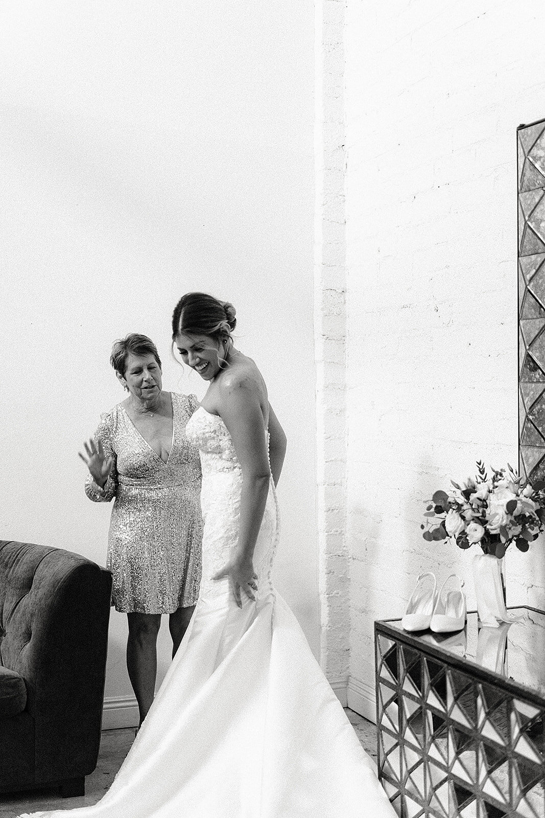 A black-and-white shot of the bride and her mother, who is wearing a sparkly dress, as they share a laugh while the bride finishes getting dressed.
