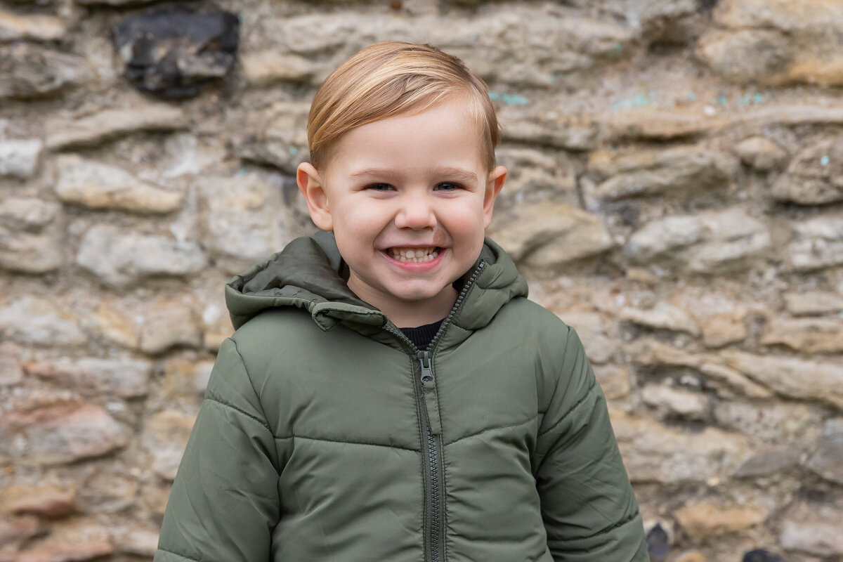 Young child smiling at the camera wearing a green jacket, standing in front of a stone wall.