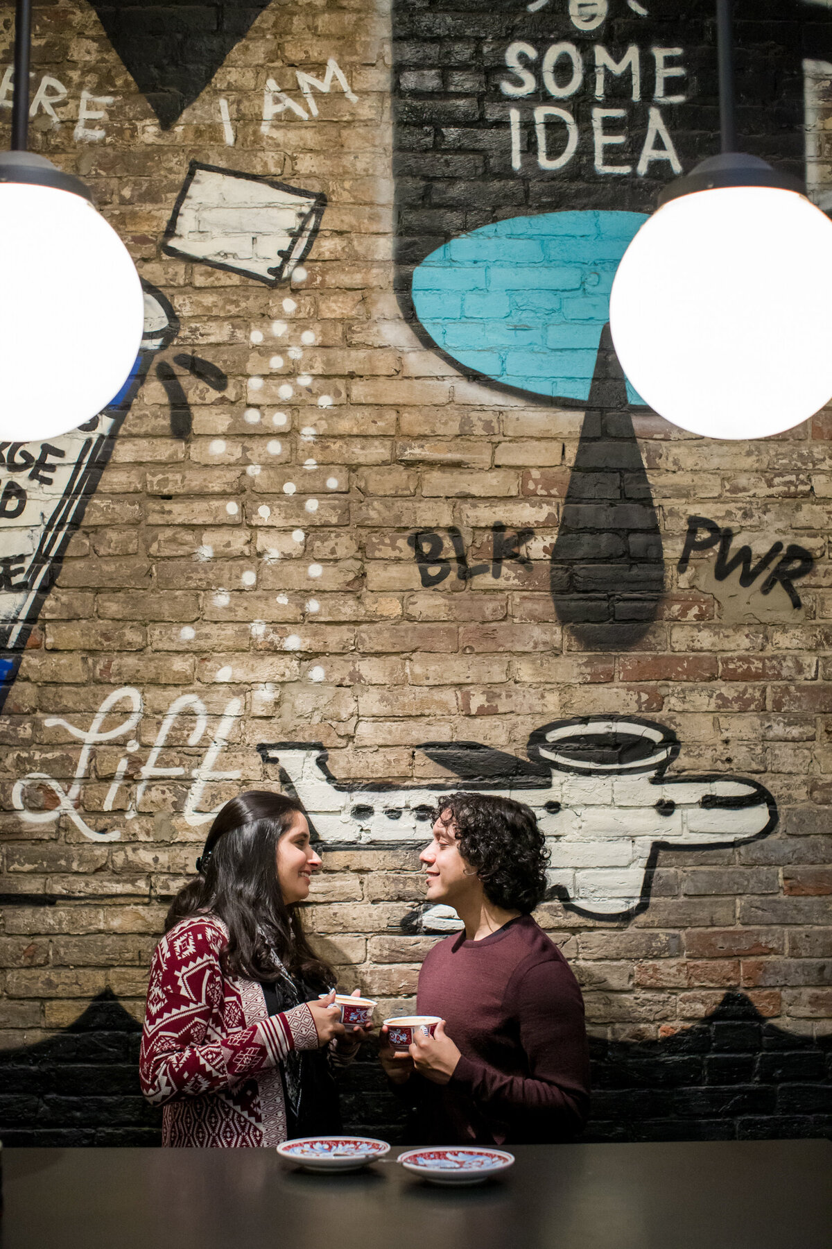 Cozy engagement photo of a couple sitting under a vibrant mural inside a Philly coffee shop, sharing a warm, artistic moment filled with personality and connection.