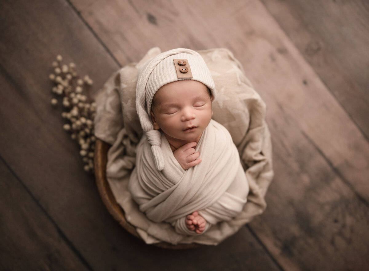 Aerial image of Lake Elsinore newborn baby photoshoot. Baby is wrapped in cream swaddle with his fingers and toes peeking out. Baby is placed in a basket and sleeping peacefully. Captured by best Lake Elsinore newborn photographer Bonny Lynn Photography
