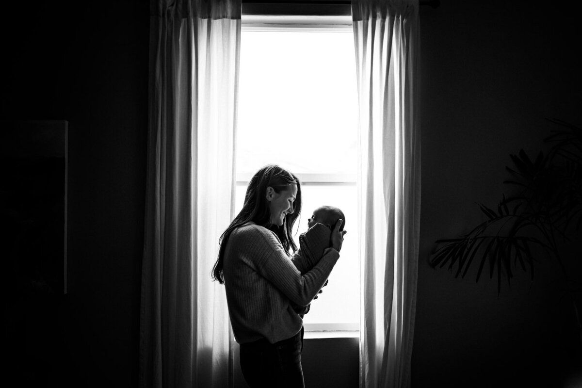 mom standing in window curtains holding newborn up to her face and smiling