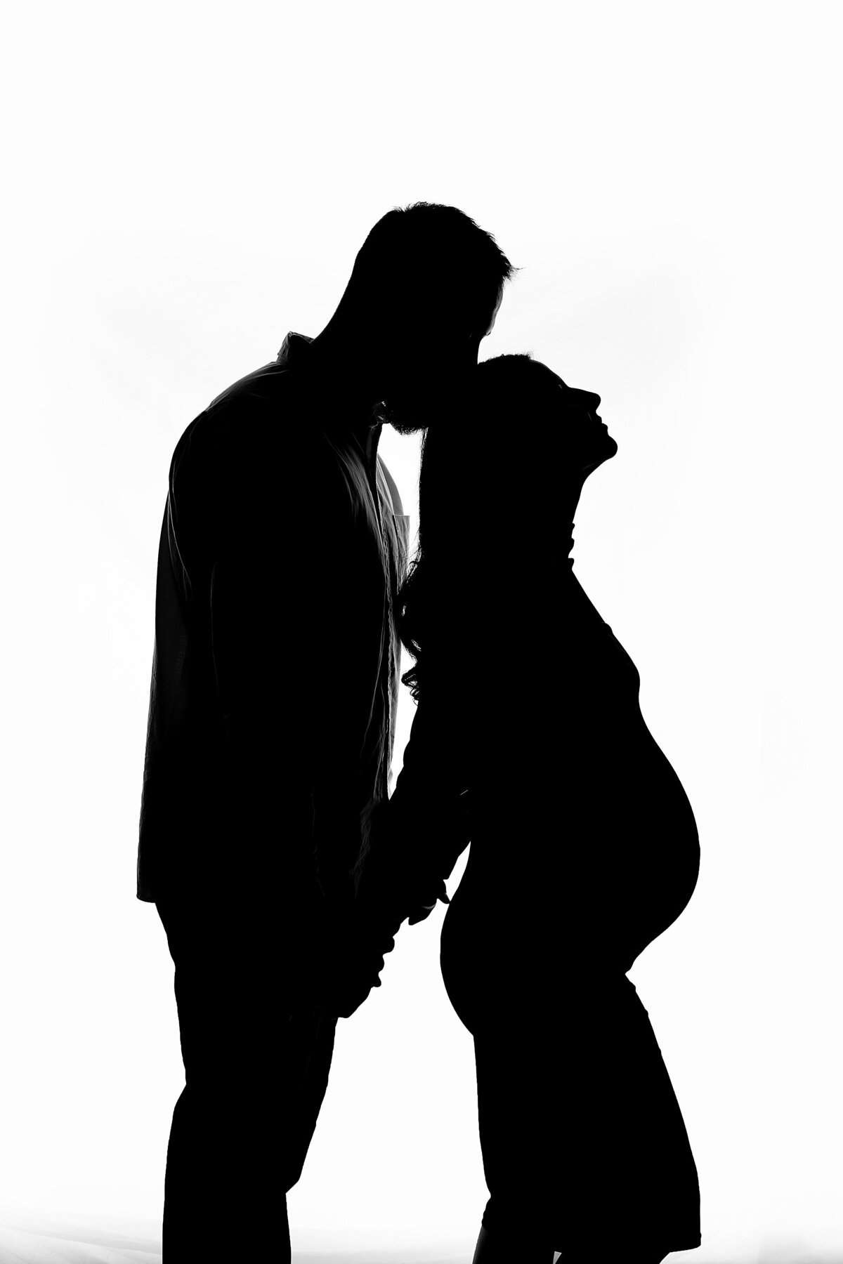 Silhouette of a couple holding hands, with the man gently kissing the pregnant woman’s forehead, captured in an emotional and artistic studio maternity session in Waukesha.