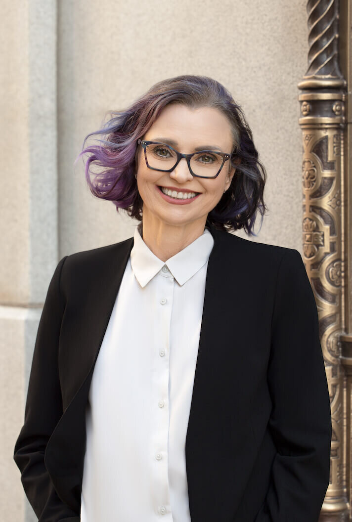 outdoor headshot of a woman standing in front a building wearing a black suit with a gray button up blouse smiling at the camera