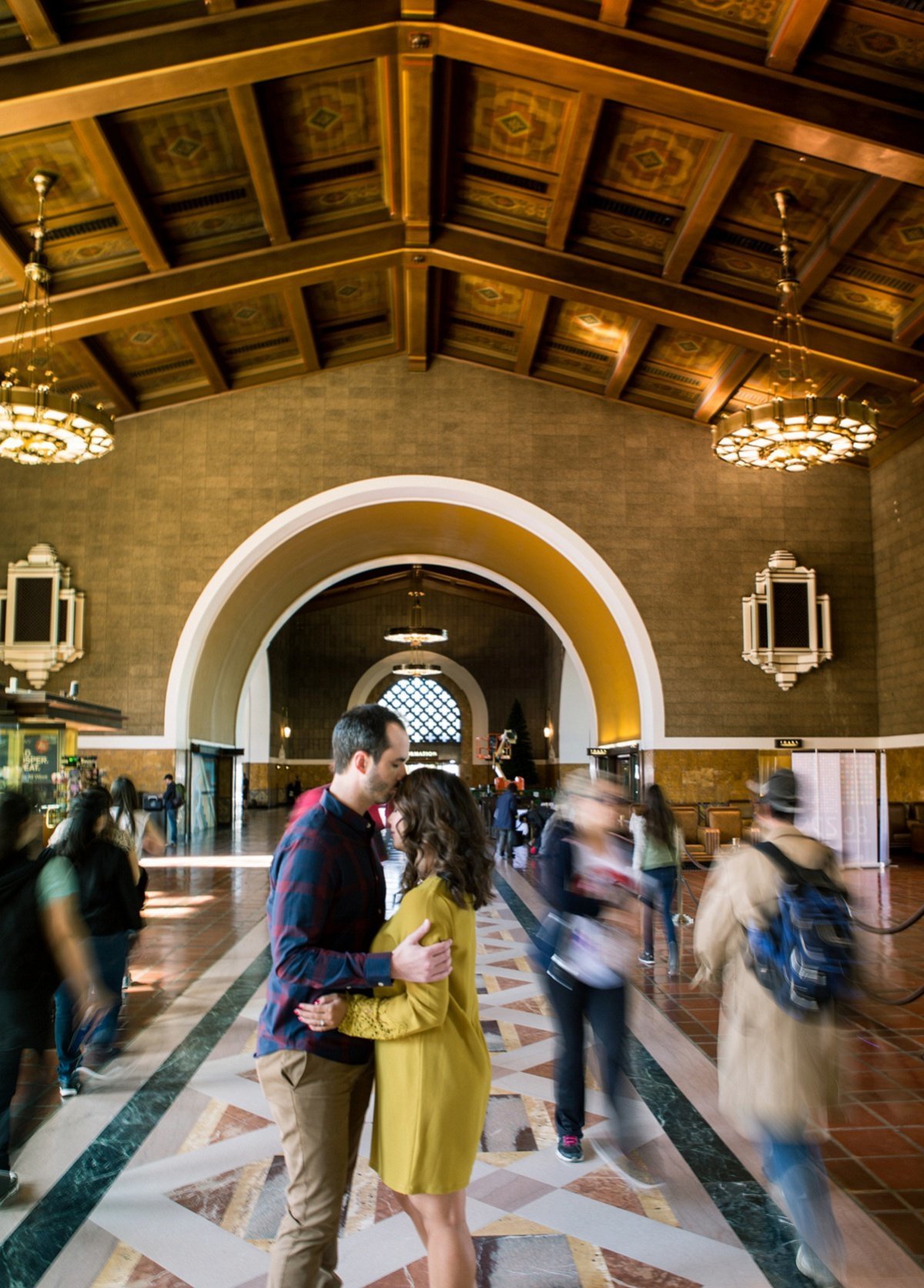 Groom to be kisses the forehead of his fiance amongst the bustle of people during an engagement photo session at the Los Angeles Union Station