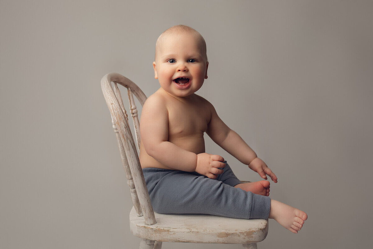 a 1 year old boy sitting on a white chair with a huge smile