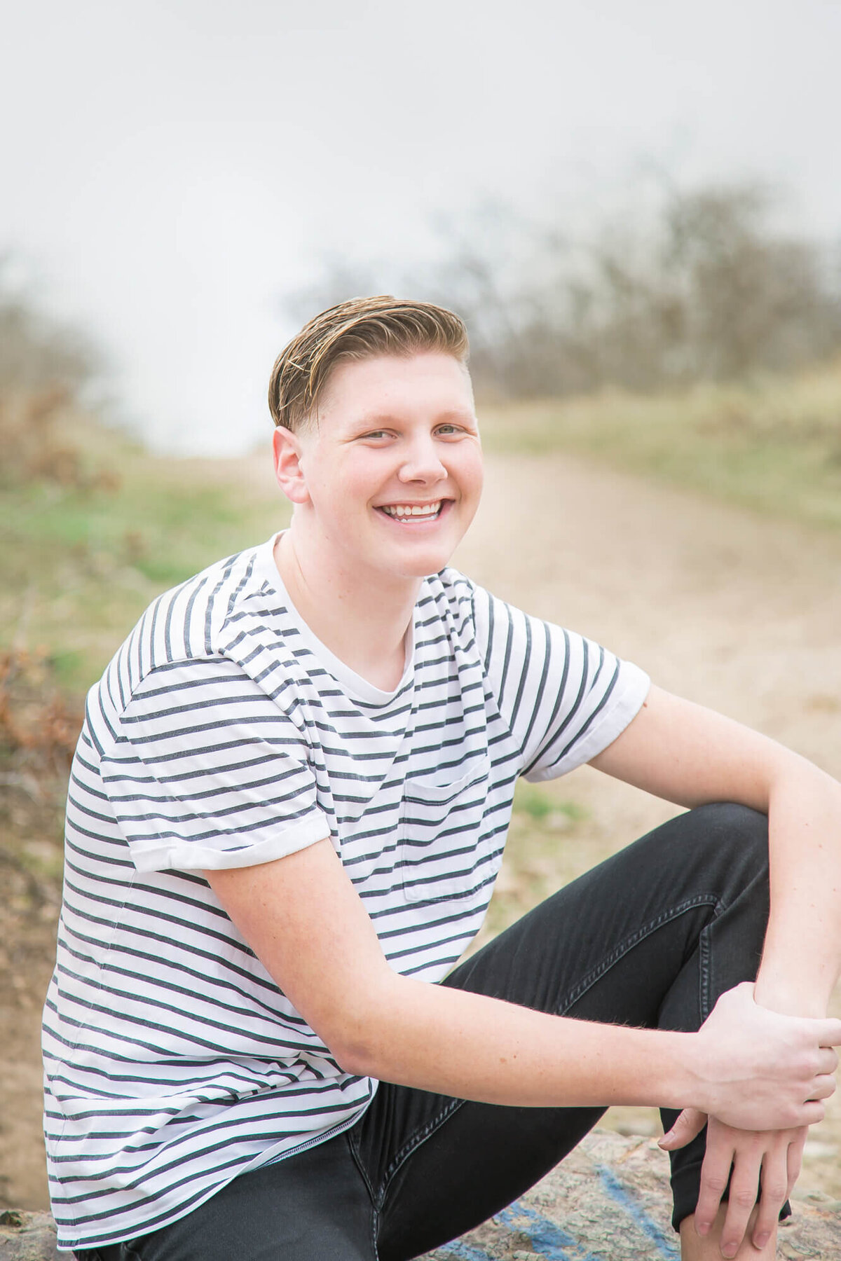 Young man  ina black and white tshirt smiling on a rock in front of a dirt path