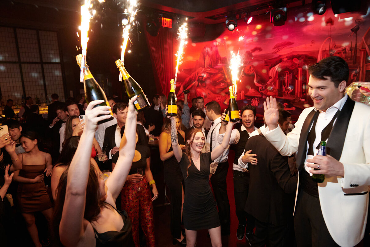 The vibrant and energetic atmosphere in the photo captured at the New York Public Library by photographer Shawn Connell, with videography by Aaron Navak Films, adds an interesting contrast to the usual serene setting of the library. The scene showcases a group of people celebrating at a nightclub, with several jubilant individuals in the foreground holding bottles of champagne adorned with sparklers. The background is bustling with more partygoers and colorful lights, creating a truly festive and lively ambiance at this unique location in New York City.