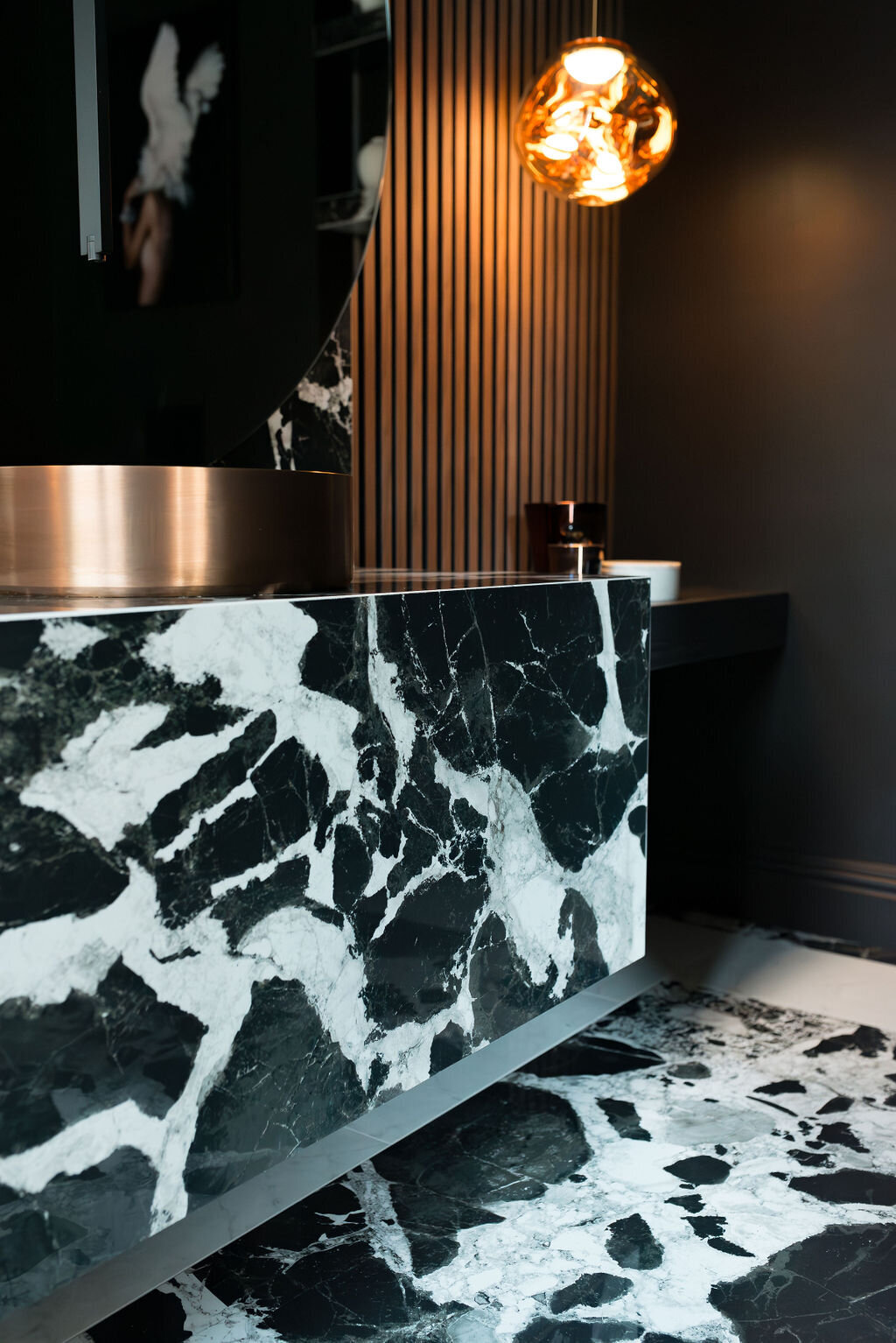 A close-up view of the marble countertop in a dark, modern bathroom. The marble extends to the flooring. The sink features a round, metallic copper bowl.