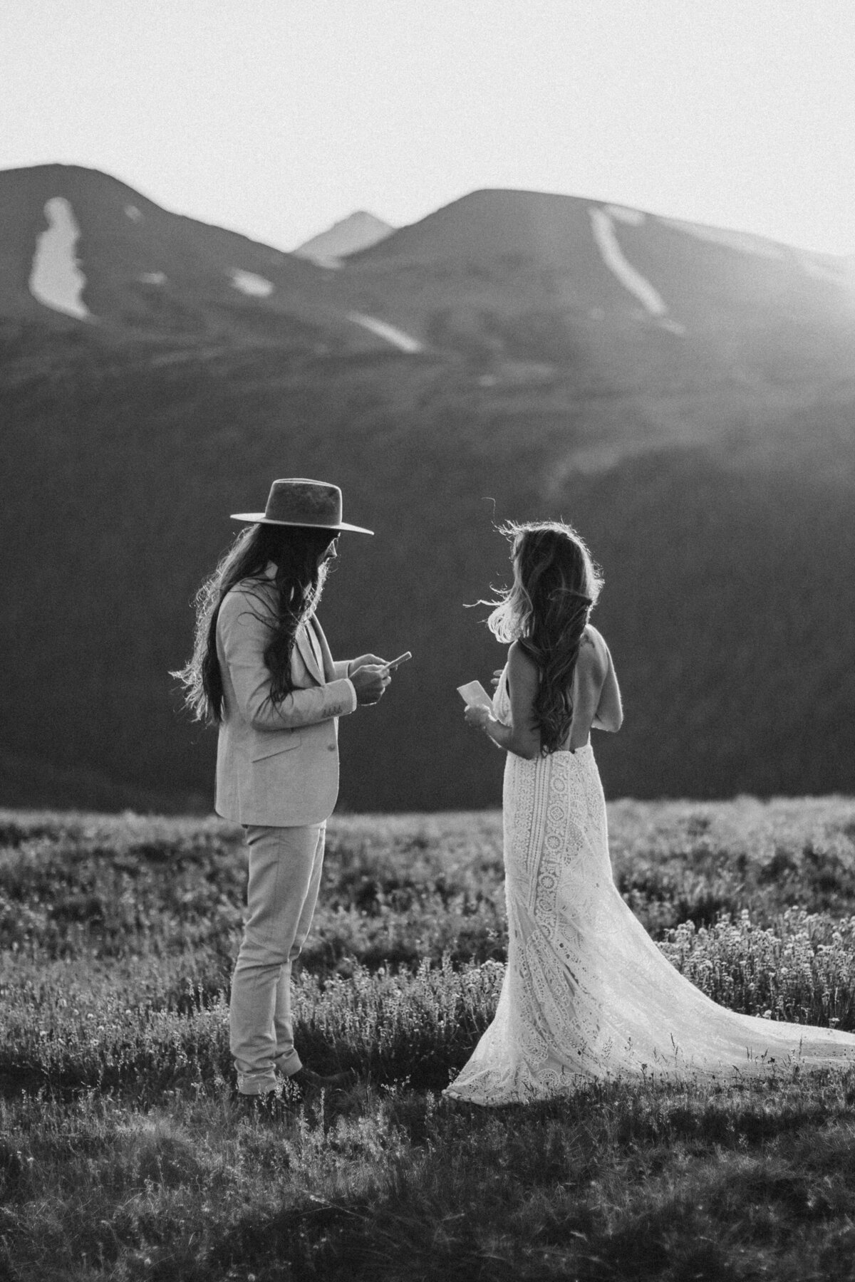 bride and groom wearing a wedding gown and tuxedo read vows  to each other in the mountains