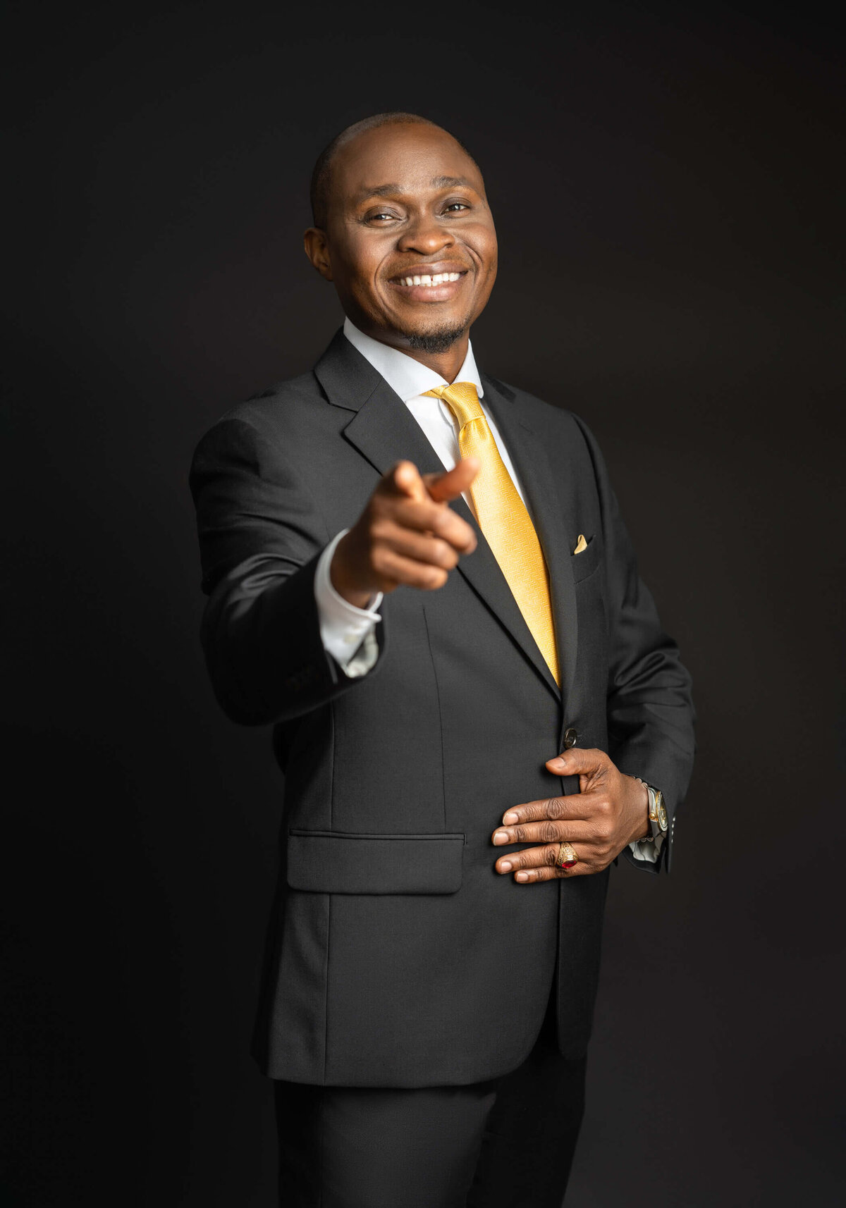 Man in a black suit with a yellow tie, pointing and smiling confidently.