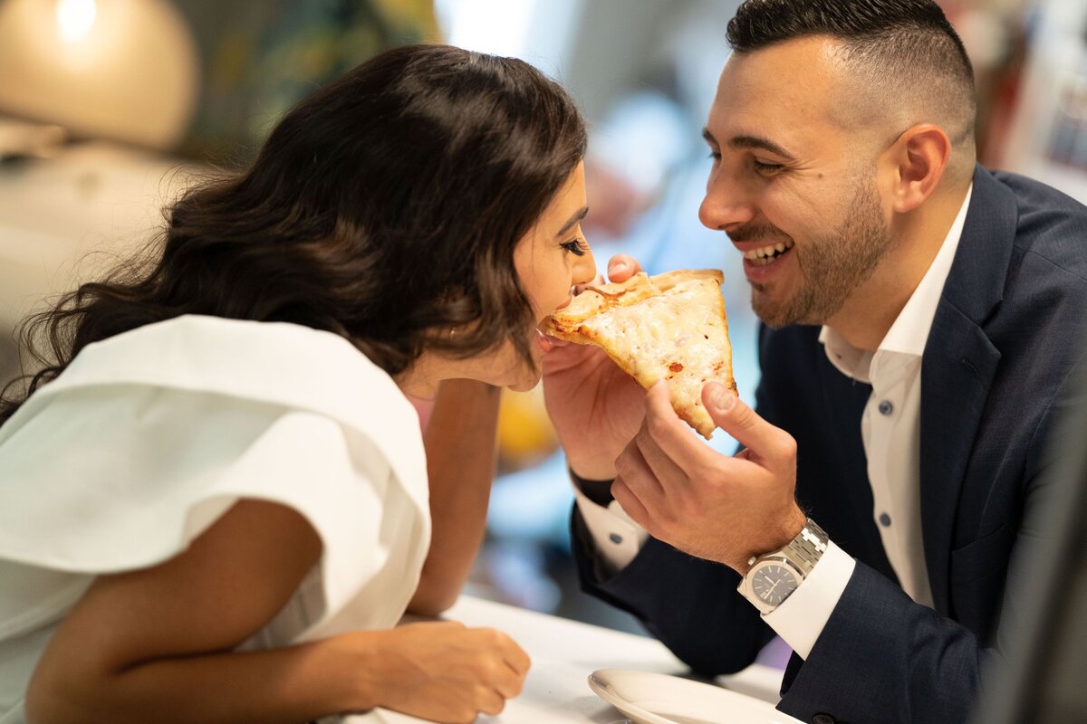 A couple joyfully shares a slice of pizza, smiling at each other. The casual and intimate moment captures their connection and the playful side of their relationship.