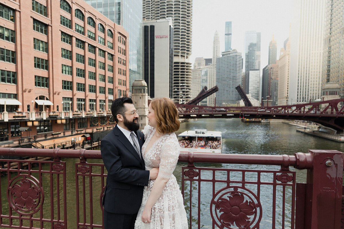 Just married couple standing very close on top of a steel bridge over the Chicago River. The view of the city is in the distance.