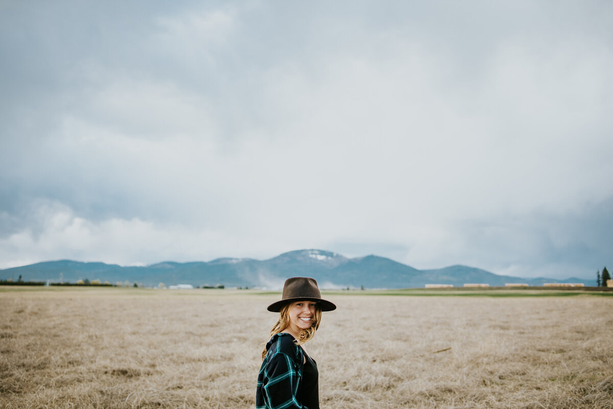 Young girl smiles at camera wearing a fedora while the remainder of the frame is filled with field, mountains, and sky.