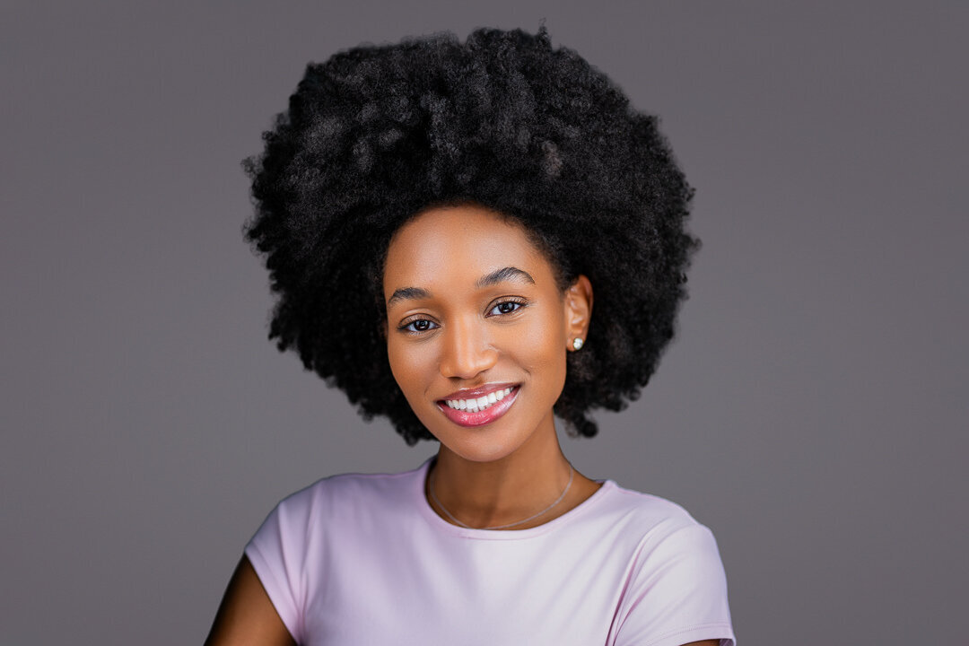 Professional model headshot in lavender top against gray background, showing natural beauty, confident smile, and clean studio lighting by Alex Kaplan Photo in NYC.