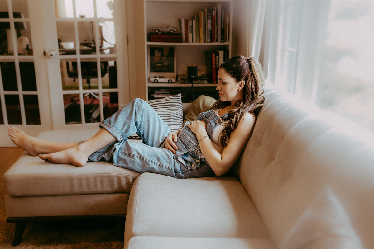 Pregnant woman in denim overalls resting on a beige sofa, holding her belly, with a bookshelf and glass-paneled doors in the background.