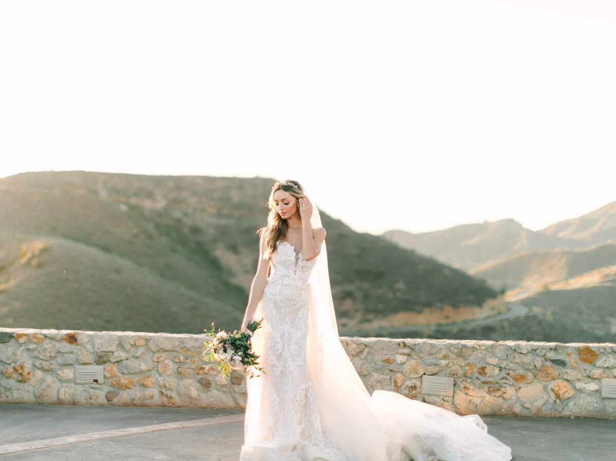 Bride in a flowing white dress stands on a terrace with mountain views, holding a bouquet.