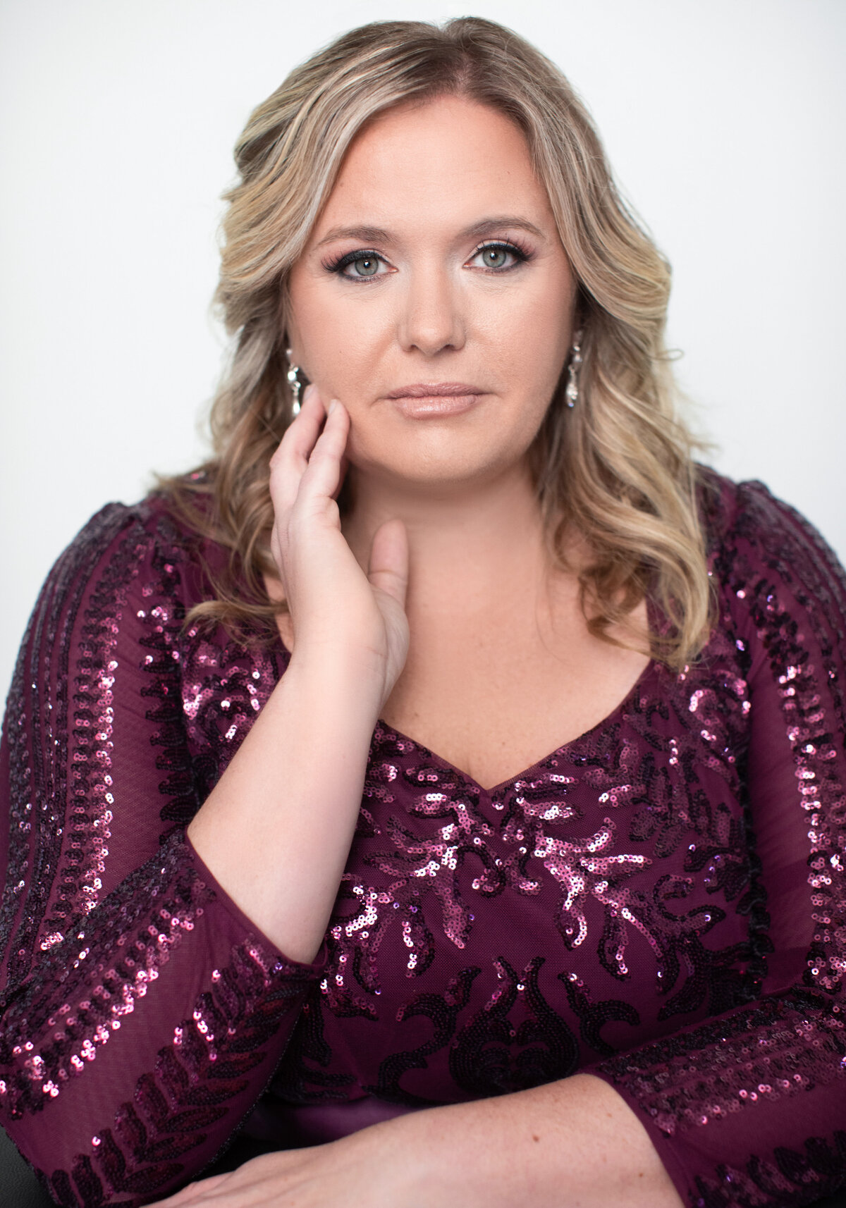 A blonde woman with red sequin dress touches her face and with an intense stare expression poses for a portrait at Janel Lee Photography studios in Cincinnati Ohio