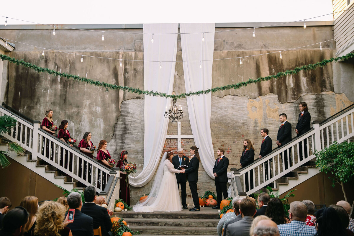 A bride and groom stand at an outdoor altar, holding hands under white drapes and a chandelier, surrounded by their wedding party on staircases. Guests are seated, and pumpkins decorate the steps.