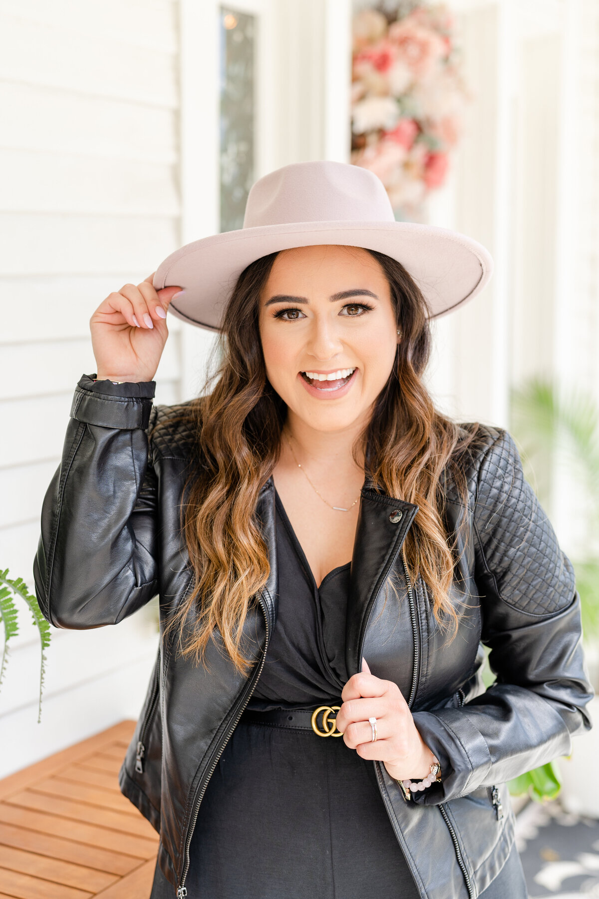 Woman laughing at camera holding black leather jacket and pink hat while standing on a front porch