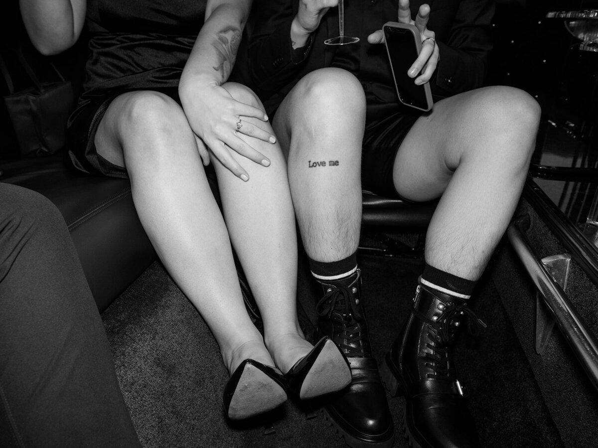 A black and white photo inside a limo of a newlywed queer couple showing off her wedding ring and their "love me" tattoo just below their knee. 
