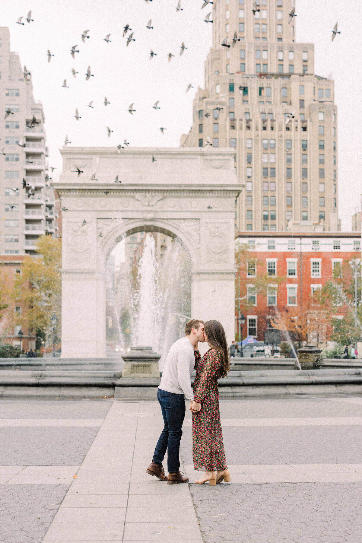 An engagement photo taken at Washington Square Park in New York City with pigeons flying overhead