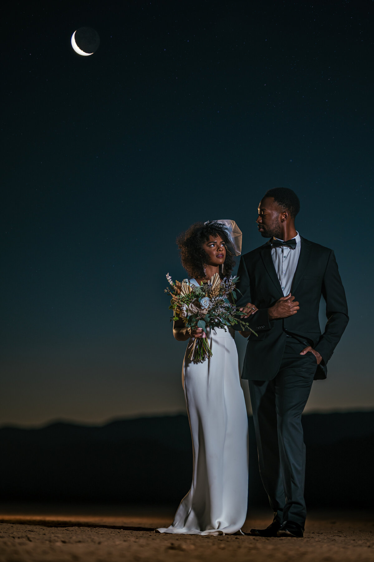 night time wedding  couple looking at each otherWedding by moonlight Dry lake Bed elopement Blue Suit on Groom  flowers by michelle  bride in cream color wedding dress with deep  plunging  neckline mountain skyline  sunset las vegas wedding photographers mk delacy photography