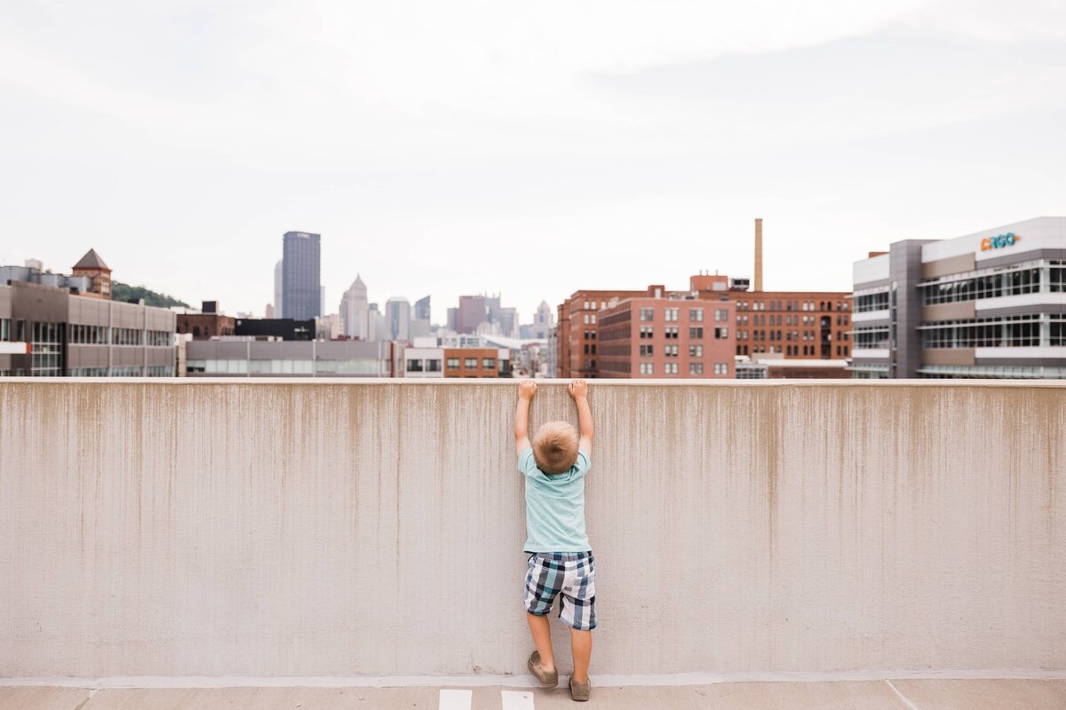 A child hangs from a wall with an urban skyline in the background, captured by a family photographer in Pittsburgh.