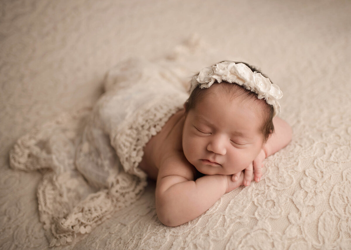 Lake Elsinore newborn photoshoot. Baby girl is sleeping on her belly with her hands folded under her chin. There is a soft lace blanket placed atop of her.