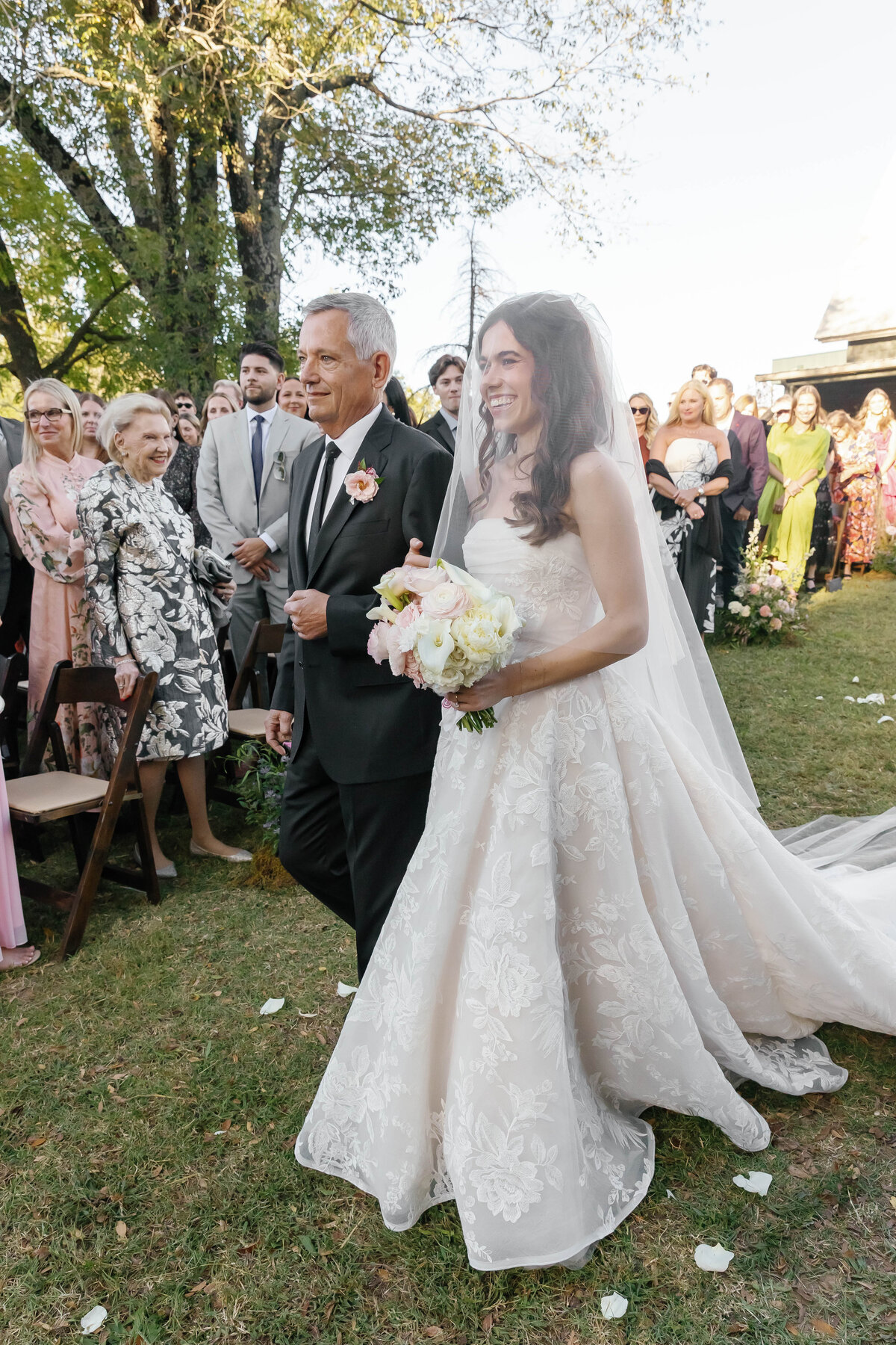 Bride walking down the aisle with her escort holding a soft pastel bouquet of roses and calla lilies during an outdoor Arkansas wedding ceremony lined with lush floral arrangements.