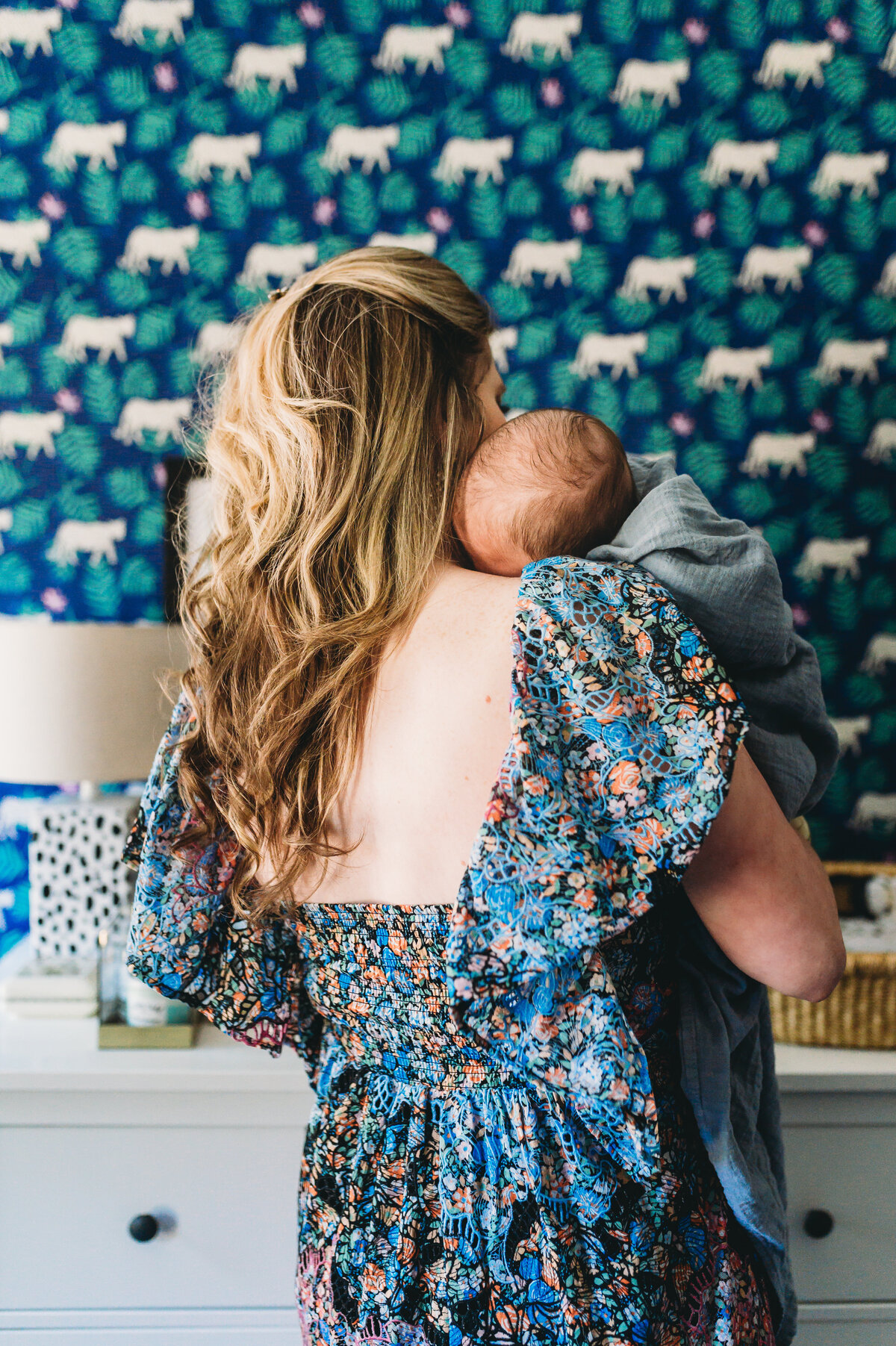 mother in floral dress holding baby boy over shoulder in nursery with wallpaler