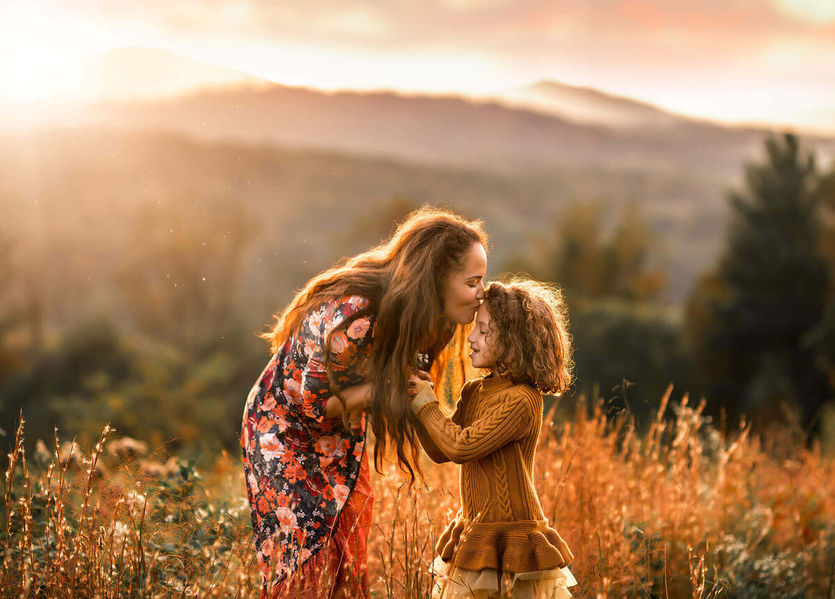 A mother kissing her daughter on the forehead with mountains in the distance at sunset