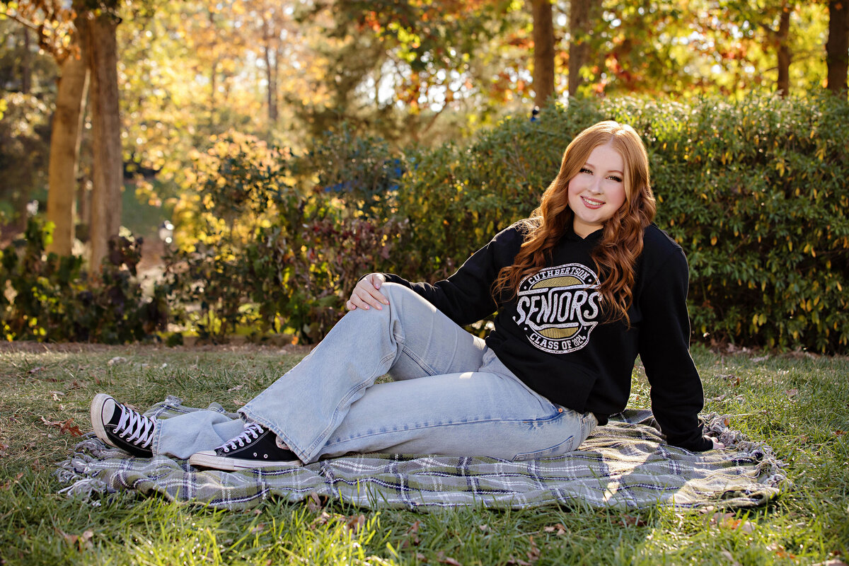 High school senior in black hoodie that reads SENIORS laying on a blanket in the grass,  and surrounded by soft fall textures