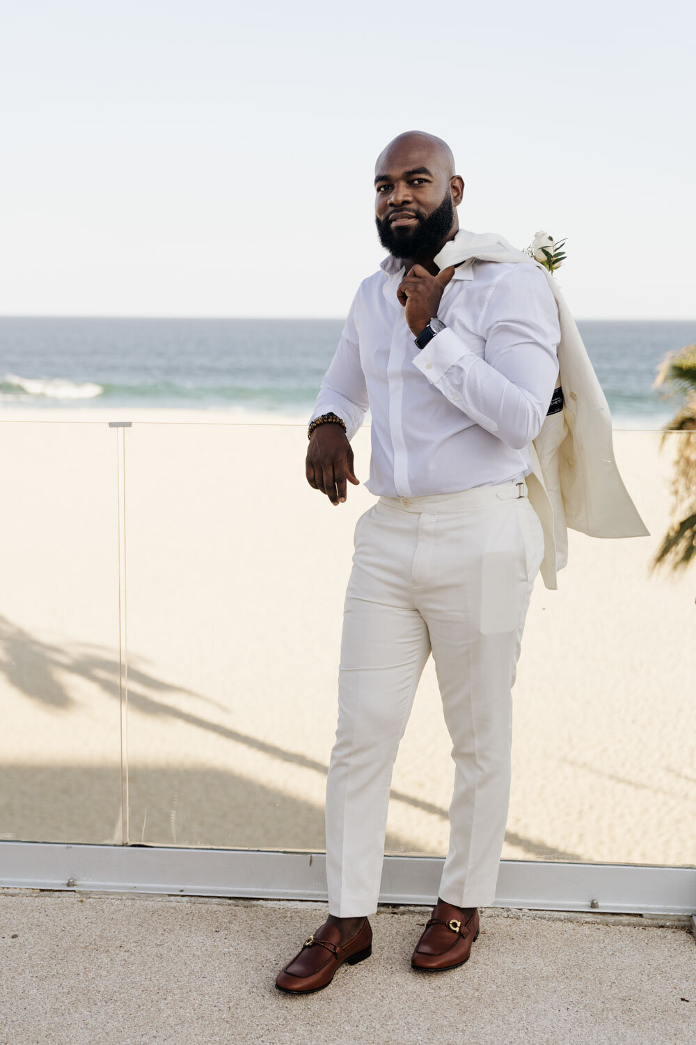 Groom in white attire standing by a beach with a jacket over his shoulder