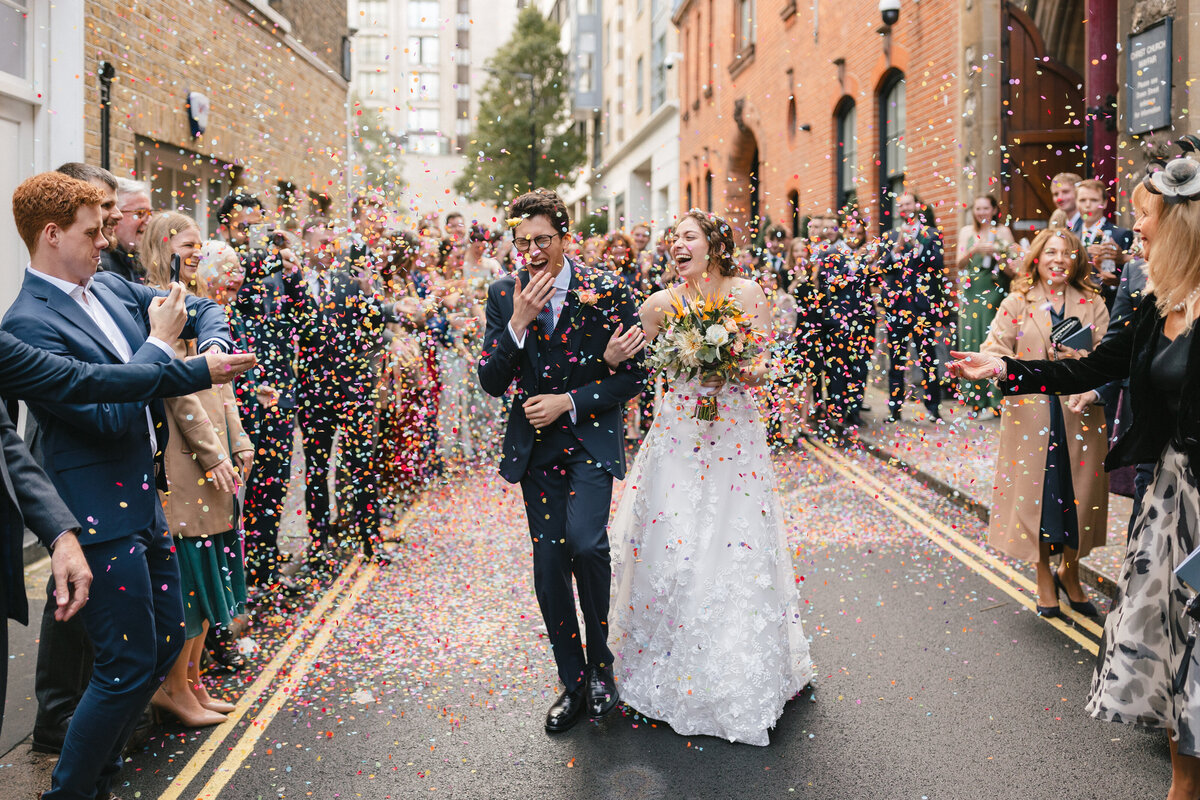 A bride and groom walk down a confetti-covered street, surrounded by cheering guests. The groom is in a dark suit, and the bride wears a white dress, holding a bouquet. It’s a joyous wedding celebration on a lively London street.