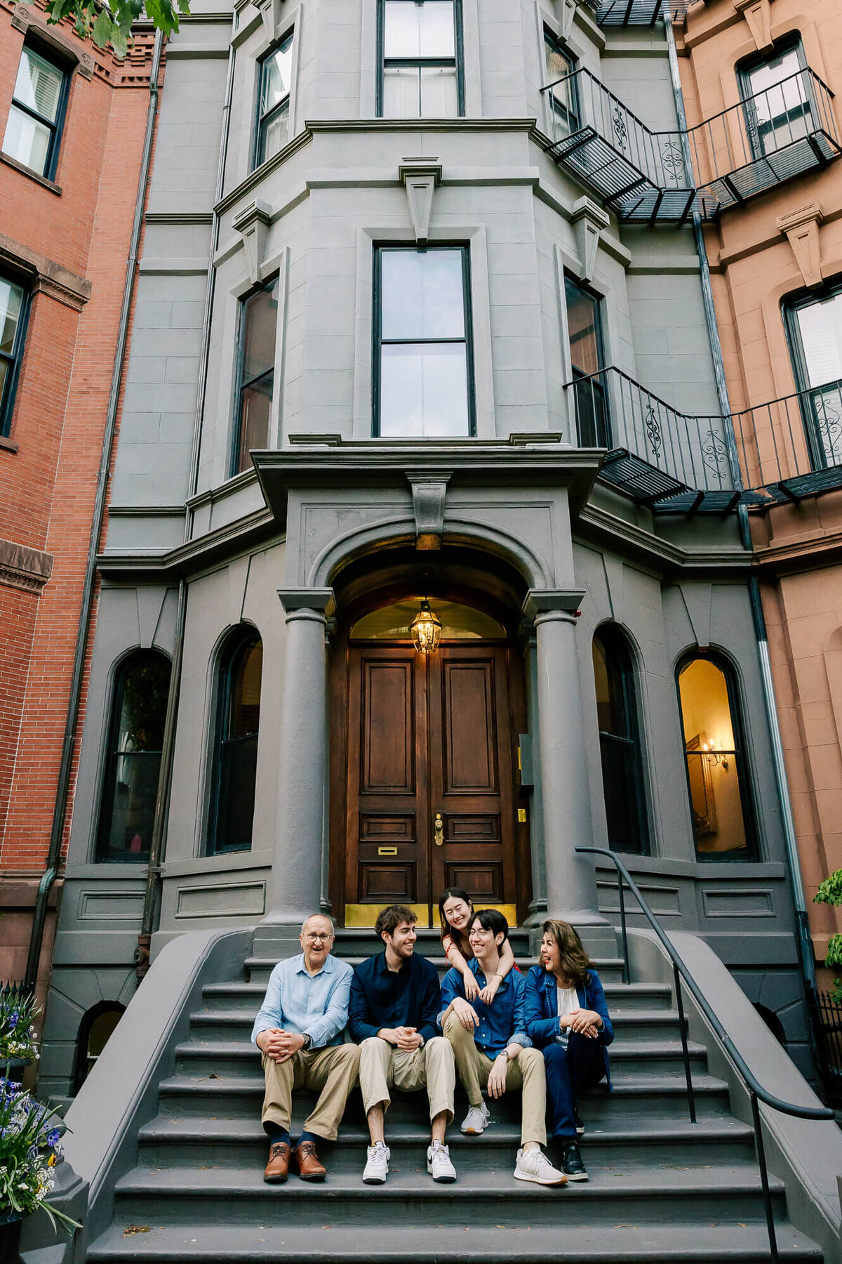 Group of 3 twenty-somethings and their two older parents, sitting on the front steps of a Boston brownstone. They're smiling and talking to each other.