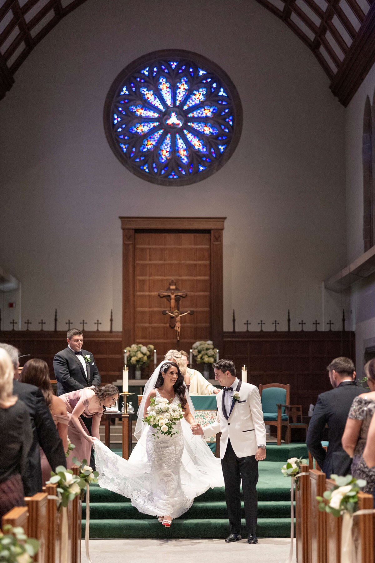 A newlywed couple walks down the aisle in a church adorned with stained glass windows. The bride holds a bouquet and wears a lace gown, while the groom wears a white tuxedo with black accents. Guests are seated, watching the couples exit.