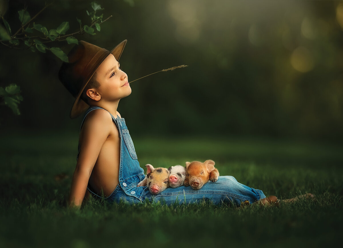 Farmer boy in cowboy hat with a straw in his mouth, wearing blue jean overalls and holding piglets.