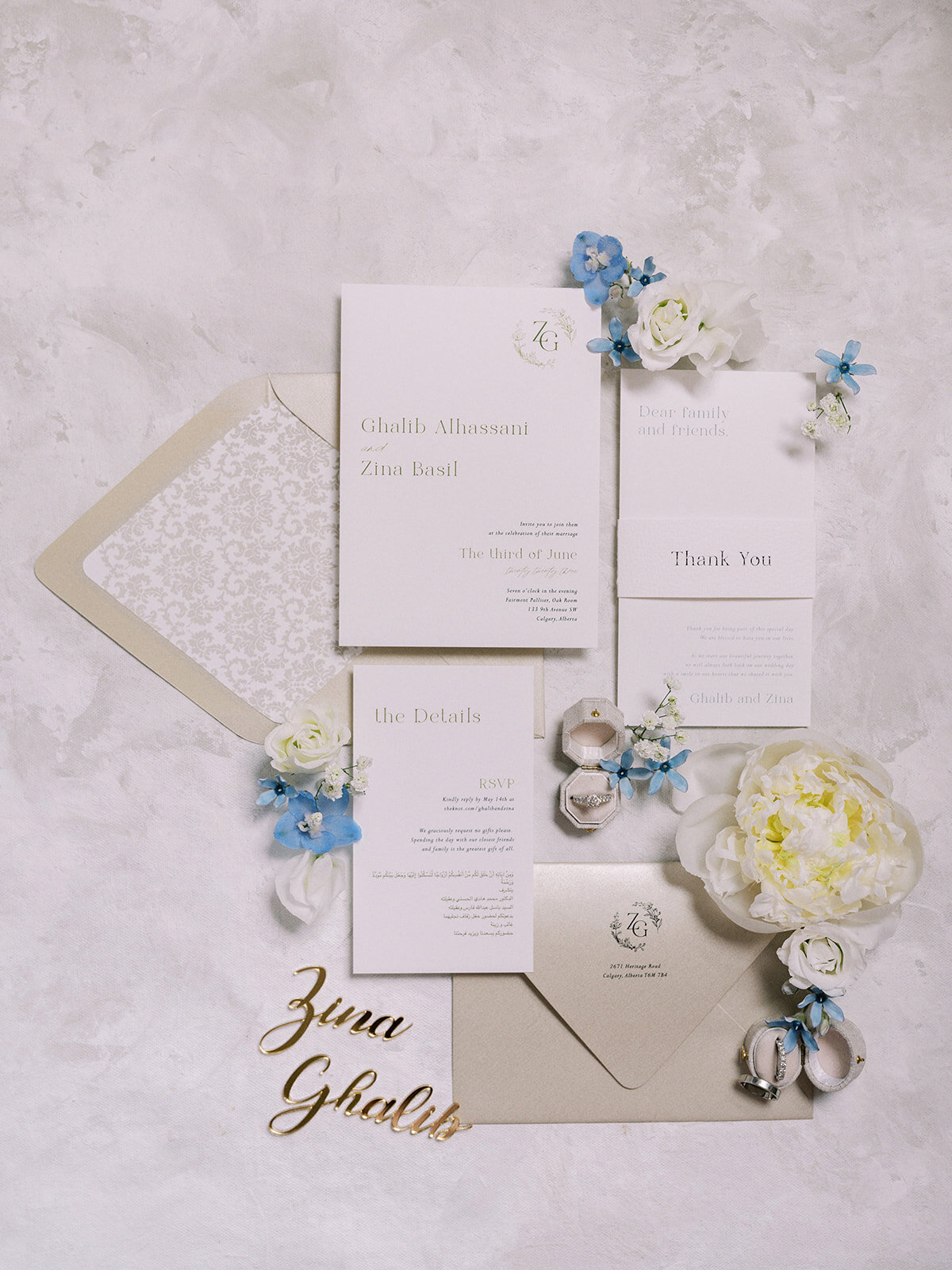 A flat lay of elegant wedding invitations and stationery for a classic Calgary wedding, including RSVP cards, a thank you card, envelopes, white flower arrangements, blue flowers, and gold name cutouts on a textured surface.