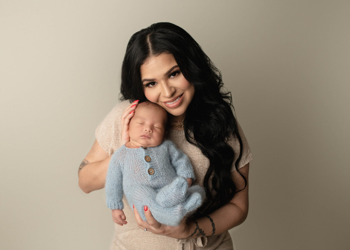 Mom poses for a photo with her newborn baby boy at his Temecula Newborn photoshoot. Baby boy is in a light blue knit onesie and mom is holding him so he is facing the camera. Mom has her cheek resting atop of the baby's head. While baby is sleeping, mom is smiling at the camera. Captured by best Temecula newborn photographer Bonny Lynn Photography