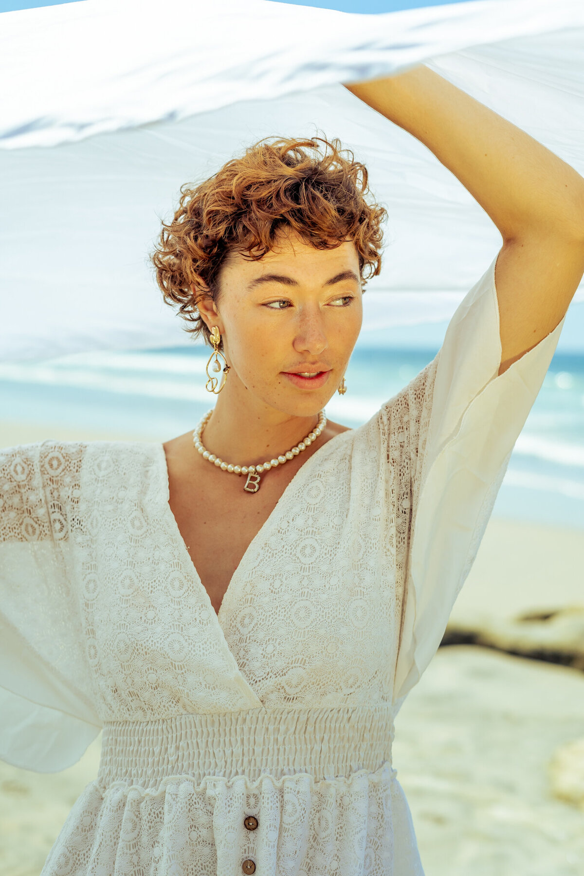 Portrait headshot of woman with short curly red hair and freckles all over light complected skin wearing a white pearl necklace and white linen dress holding a white cloth above her head blowing in the wind standing on the beach