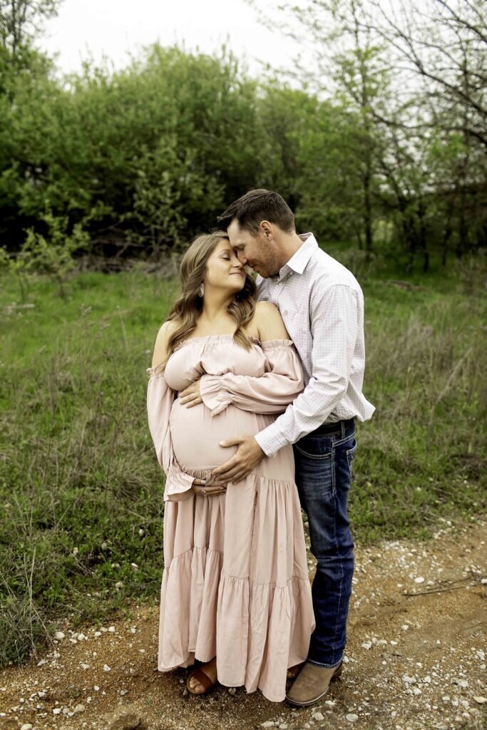 A romantic maternity photo of the expectant mom and dad standing close together in the park, with the dad gently holding his wife’s belly, both sharing a loving smile, captured by Chunky Monkey Photography.